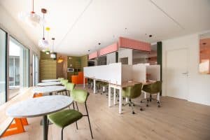 Un espace de coworking moderne avec des sols en bois clair, des chaises vertes, des tables en marbre blanc et des postes de travail divisés par des cloisons blanches. Des lampes suspendues sont accrochées au plafond et de grandes fenêtres laissent entrer la lumière naturelle.