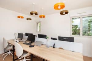 Un bureau en open space moderne comprenant six postes de travail, chacun équipé d'écrans, d'ordinateurs portables et de lampes de bureau. La pièce présente des murs blancs, une grande fenêtre et des luminaires de plafond originaux rehaussés de touches orange et dorées.